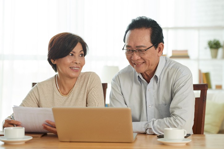 Elderly couple sitting at a table signing up for Medicare with a laptop, shopping for Medicare plans online.