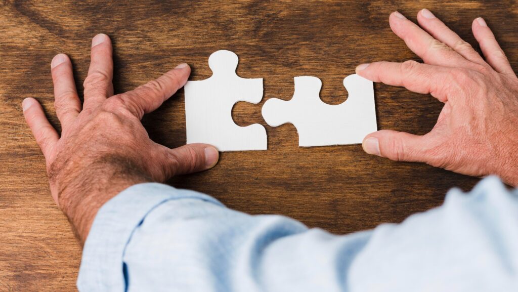 Senior hands fitting two white puzzle pieces together on a wooden table